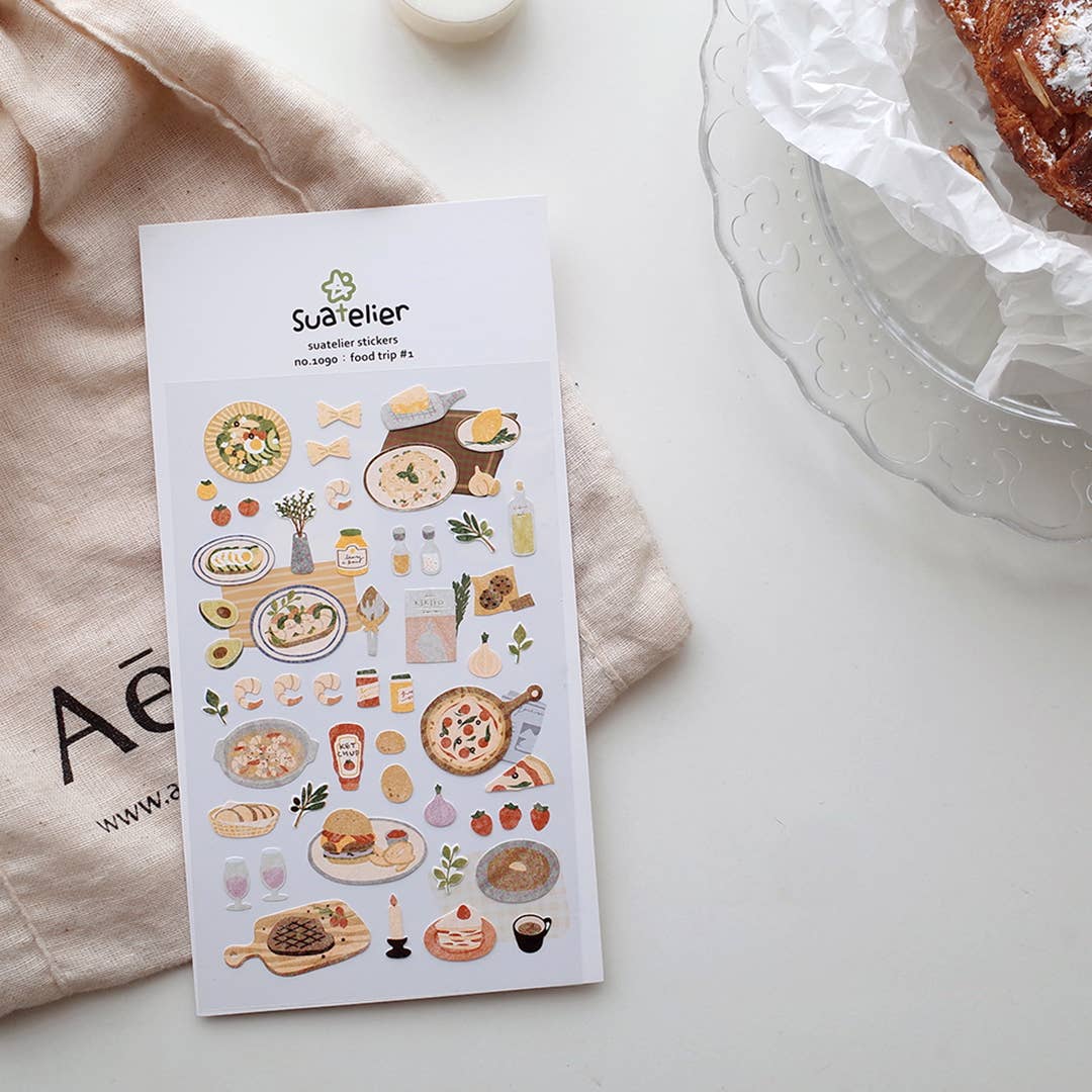 Sheet of food-themed stickers by Suatelier on a white surface with a beige cloth and glass container in the background.