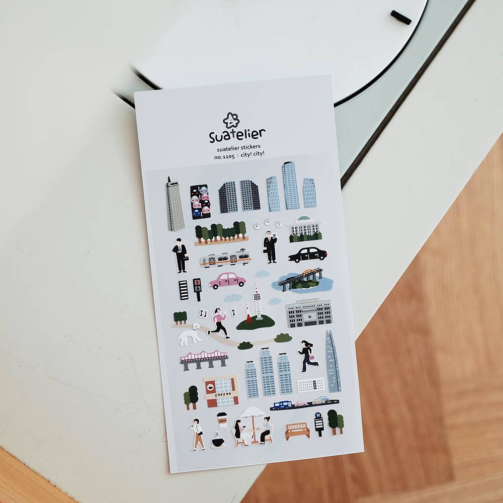 Sheet of city-themed Suatelier stickers on a white surface with a clock in the background.